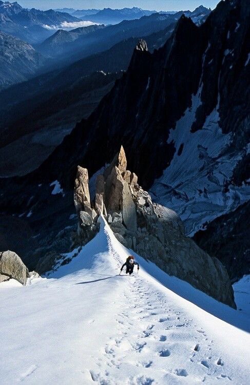 Ein Bergsteiger erklimmt einen schneebedeckten Gipfel, umgeben von hoch aufragenden Felsen und schneebedeckten Bergen im Hintergrund.