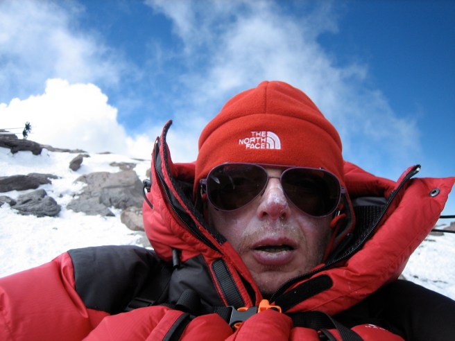 Person in roter Winterjacke und Mütze auf einem schneebedeckten Berggipfel, mit Sonnenbrille, umgeben von Wolken.